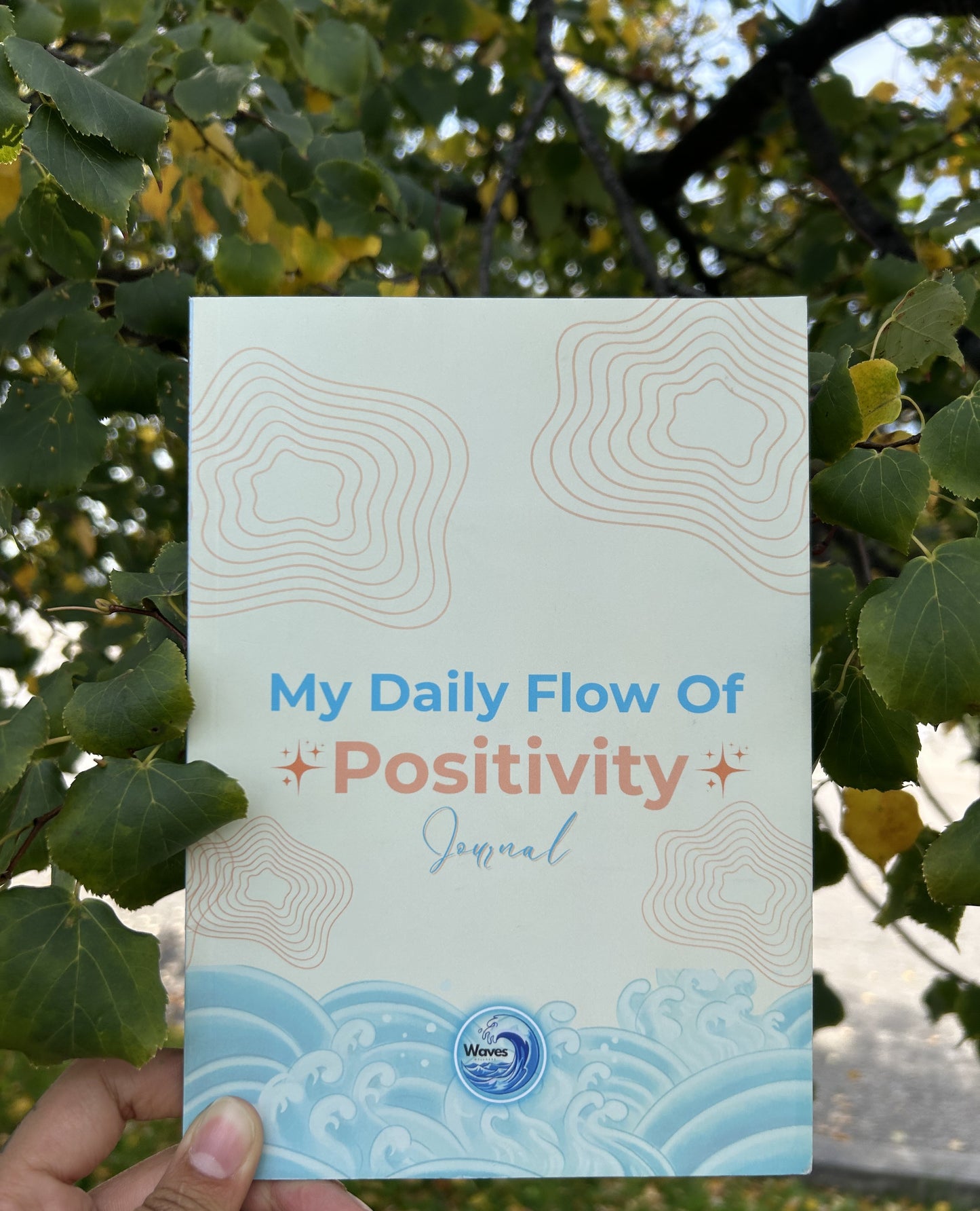 Journal titled 'My Daily Flow Of Positivity' held in front of green foliage